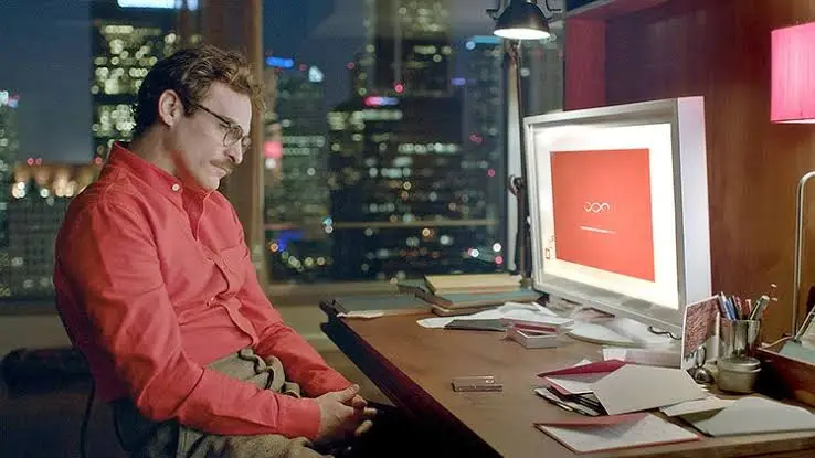 Cena do filme Her: Theodore à mesa, ao lado do computador com a interface do sistema operacional.