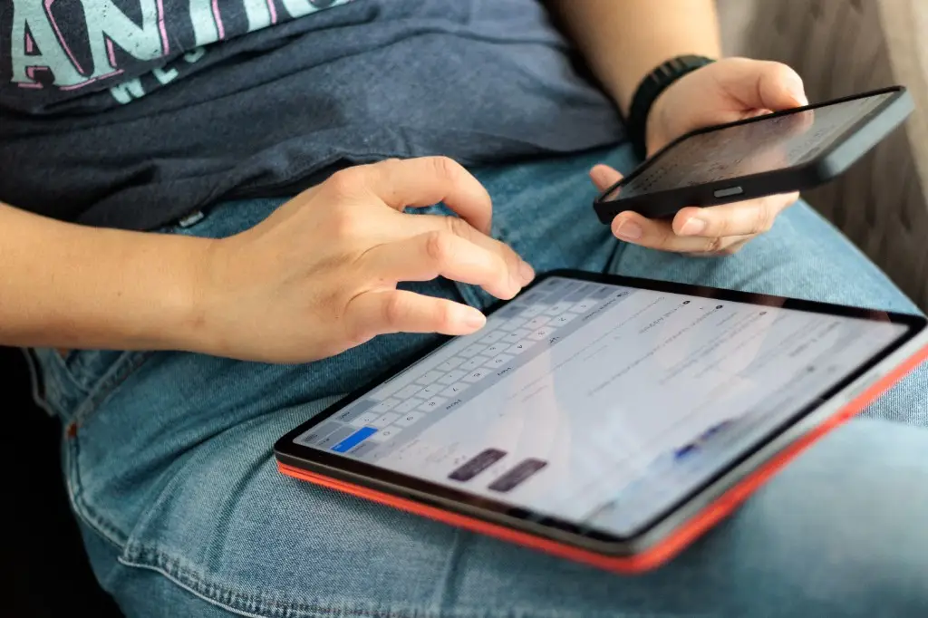 Pessoa usando um tablet no colo e segurando um celular, com teclado na tela.